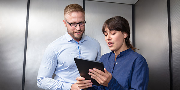 Two professionals standing in an elevator while a woman presents an idea on a tablet to her colleague.